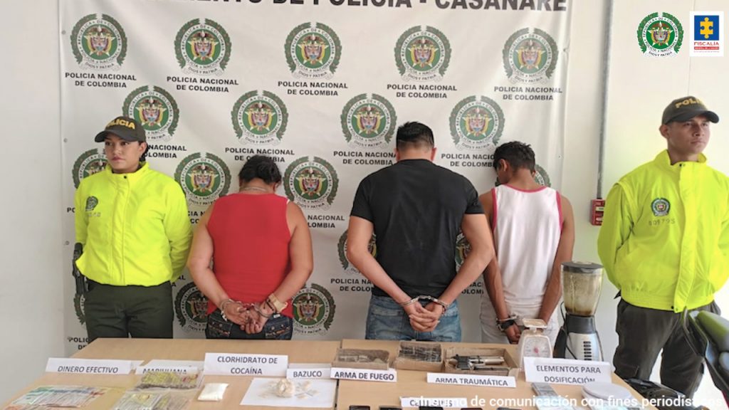 los capturados de espaldas custodiados por dos uniformados de la Policía, delante de ellos una mesa con material incautado, detrás banner de la Policía