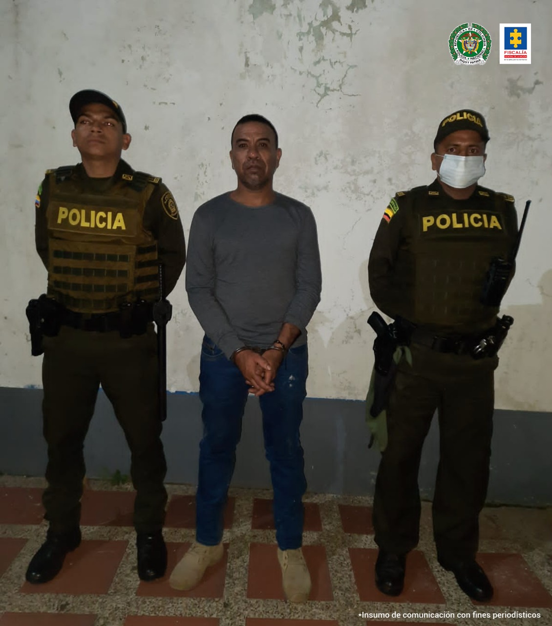 El capturado tiene puestas las esposas en sus manos que están hacia adelante y está rodeado de dos uniformados de lam Policía Nacional.