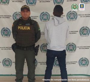 En la imagen se ve una persona privada de la libertad, de espaldas, bajo custodia de un integrante de la Policía Nacional. Al fondo un backing de la institución.