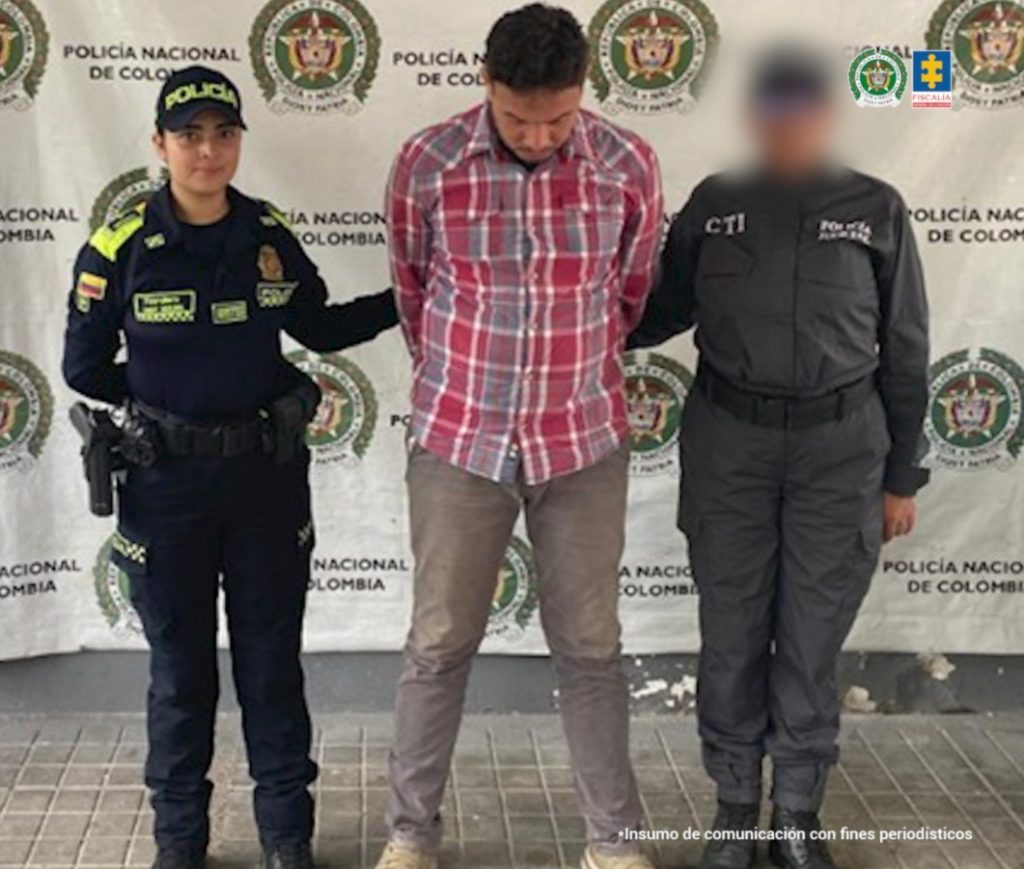 en la imagen se ve un hombre detenido bajo custodia de un investigador del CTI y un integrante de la Policía Nacional. Detrás suyo un backing institucional.