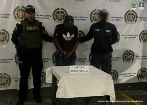 en la imagen se ve un hombre detenido bajo custodia de un investigador del CTI y un uniformado de la Policía Nacional. Detrás suyo un backing institucional.