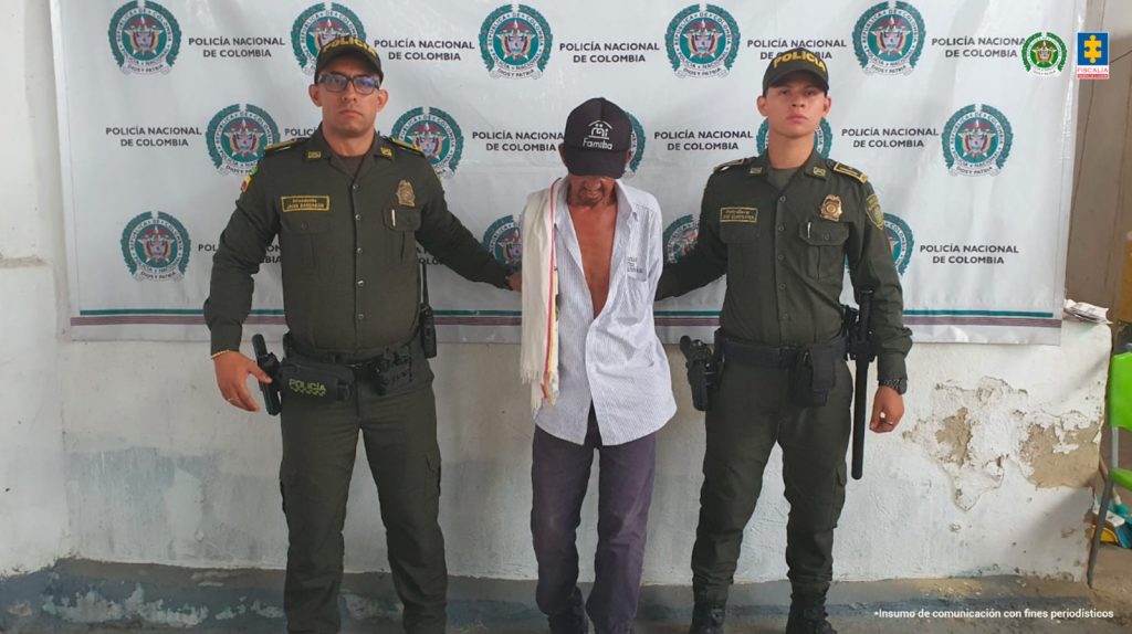 En la imagen se ve un hombre detenido bajo custodia de dos uniformados de la Policía Nacional. Detrás suyo un backing institucional.