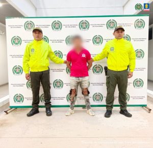 en la imagen se ve una persona detenida bajo custodia de dos uniformados de la Policía Nacional. Detrás suyo un backing institucional.