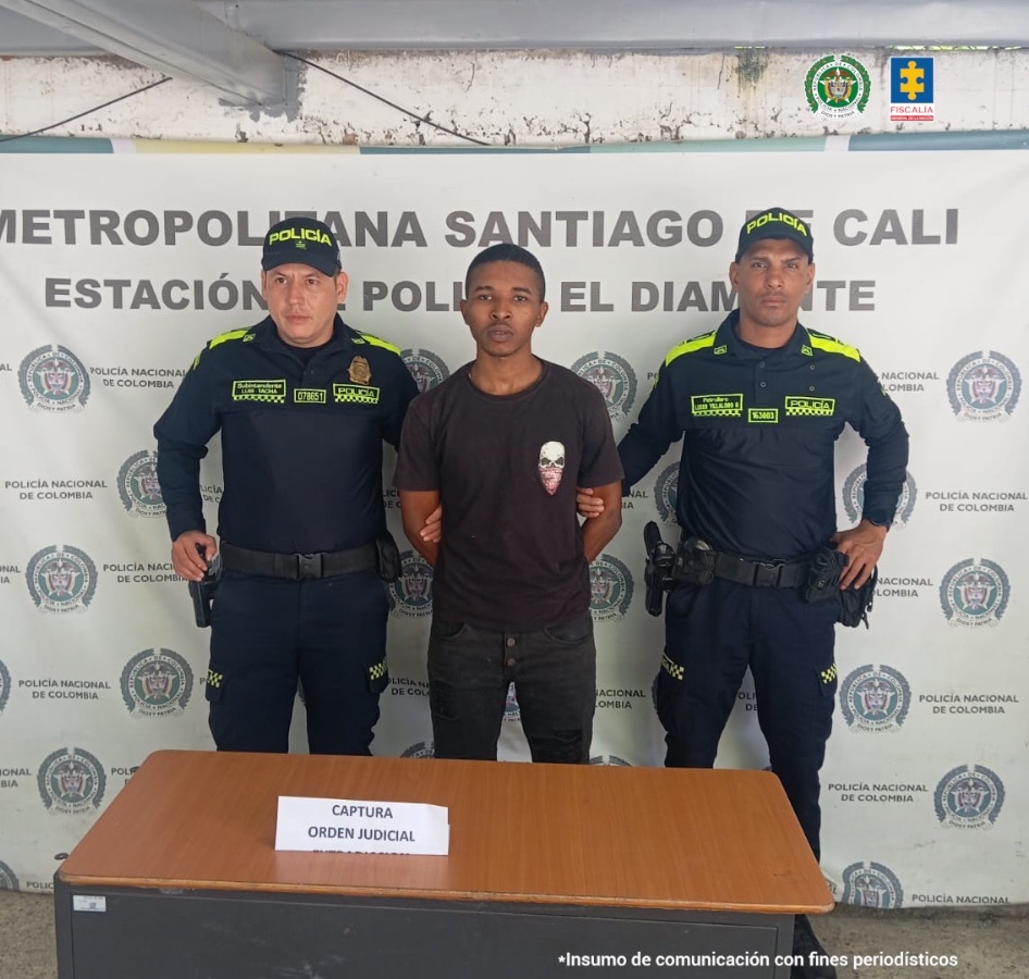 En la imagen se ve una persona detenida bajo custodia de dos integrantes de la Policía Nacional. Detrás suyo un backing institucional.