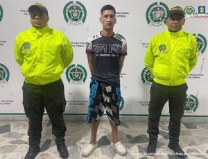 En la imagen se ve una persona detenida bajo custodia de dos integrantes de la Policía Nacional. Detrás suyo un backing institucional.