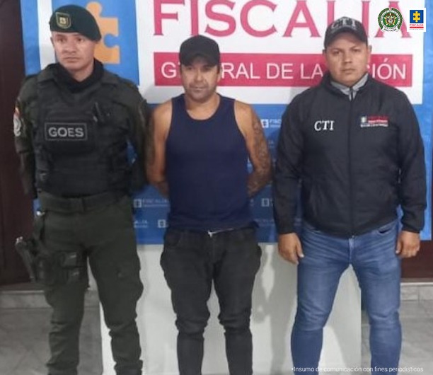 en la imagen se ve un miembro de la Policía Nacional y otro del CTI custodiando un hombre detenido. Detrás de ellos un backing de la Fiscalía.