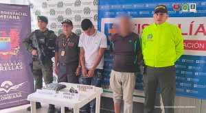 En la imagen se ve una persona detenida bajo custodia de uniformados de la Policía y el CTI de la Fiscalía. Delante suyo una mesa con elementos incautados y detrás de ellos backings institucionales.