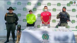 En la imagen se ve una persona detenida bajo custodia de integrantes de la Policía Nacional. Delante suyo una mesa con elementos incautados. Detrás un backing institucional.