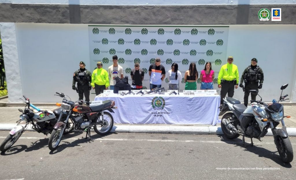 en la imagen se ven seis personas detenidas bajo custodia de cuatro integrantes de la Policía Nacional. Delante de ellos una mesa con sustancias y varios elementos incautados. Detrás de ellos un backing institucional.