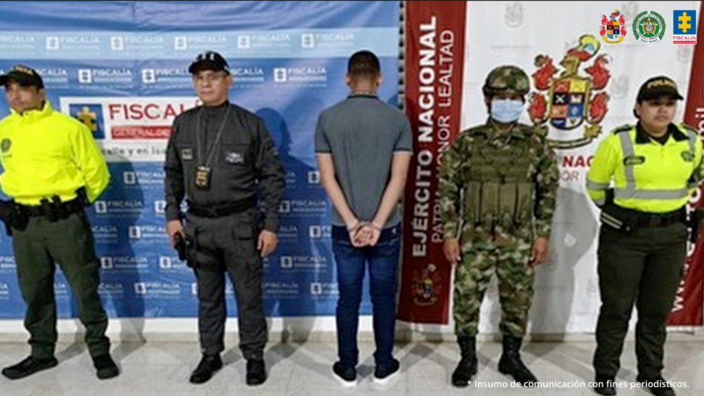 en la imagen se ve una persona de espaldas detenida bajo custodia de servidores del CTI de la Fiscalía, Policía y Ejército. Delante suyo, backings institucionales.