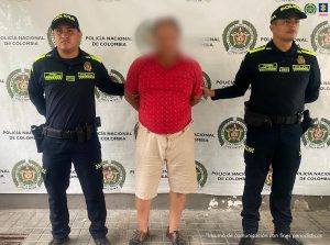 en la imagen se ve una persona detenida bajo custodia de dos uniformados de la Policía Nacional. Detrás suyo un backing institucional.