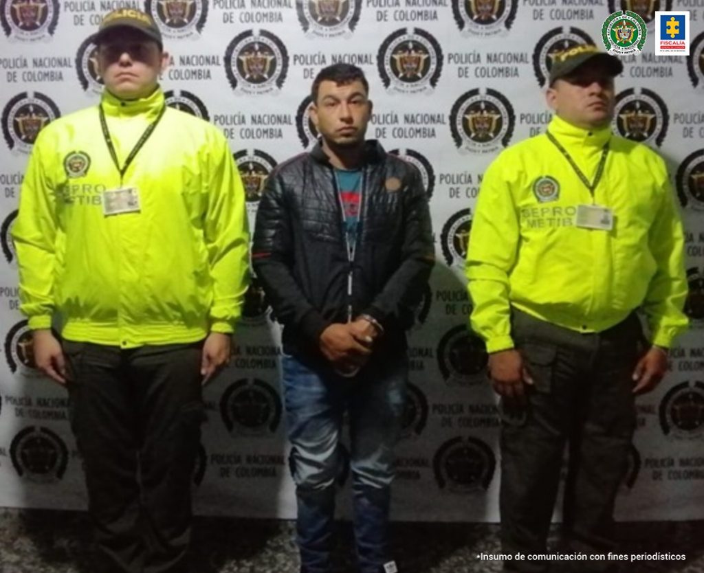 En la imagen se ve a un hombre detenido bajo la custodia de dos integrantes de la Policía Nacional. Detrás suyo un backing de la misma entidad.