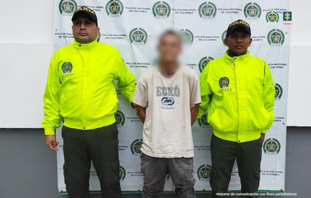 En la imagen se ve una persona detenida bajo custodia de dos integrantes de la Policía Nacional. Detrás suyo un backing institucional.