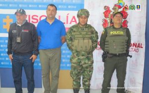 En la imagen se ve una persona detenida bajo custodia de un investigador del CTI, un funcionario de la Policía Nacional y del Ejército. Detrás suyo backings alusivos a las instituciones.