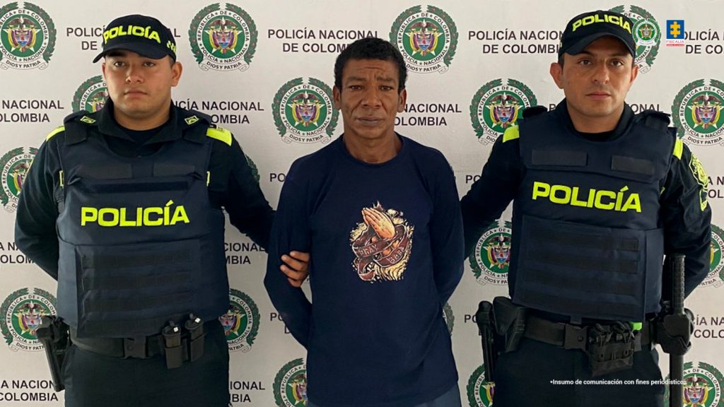 En la imagen se ve un hombre detenido bajo custodia de dos integrantes de la Policía Nacional. Detrás suyo, un backing institucional.