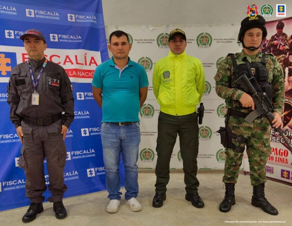 En la imagen una persona detenida bajo la custodia de un investigador del CTI, un miembro del Ejército y de la Policía Nacional. Detrás suyo un backing de la Fiscalía General de la Nación, de la Policía y el Ejército.