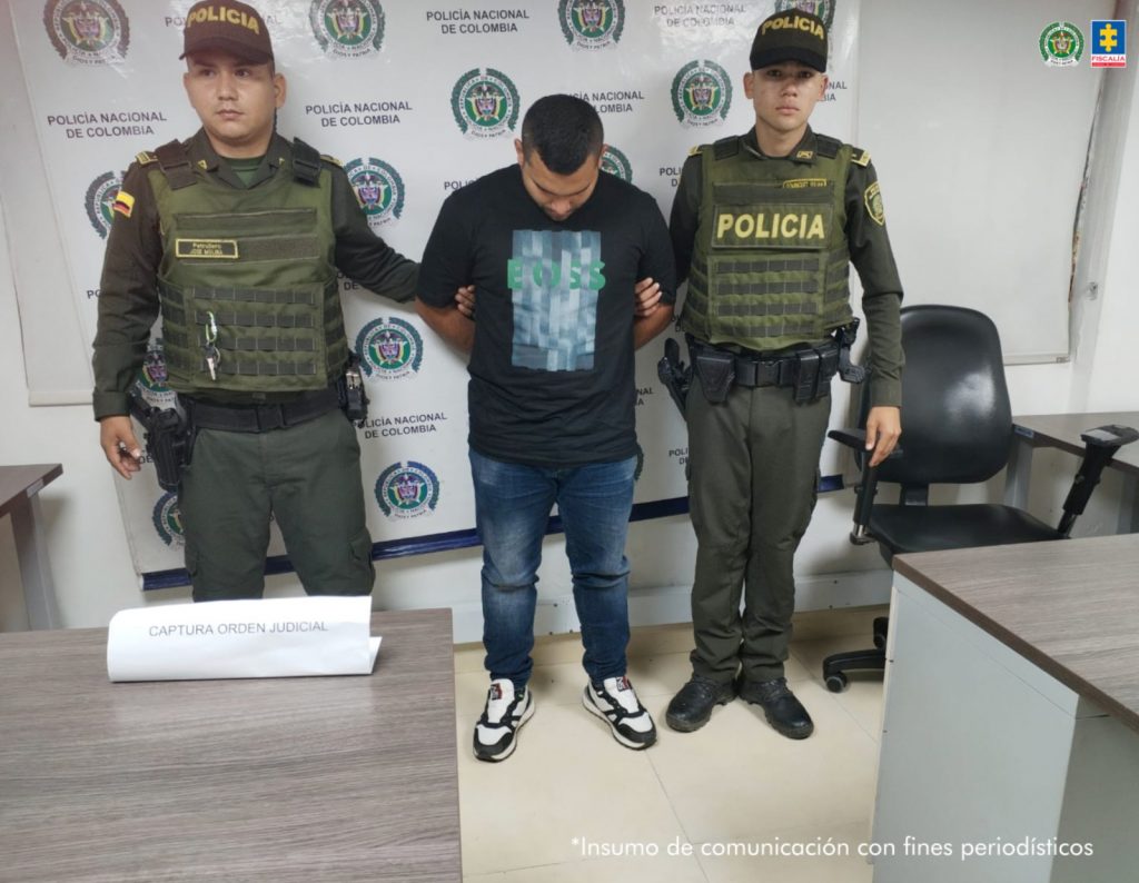En la imagen se ve una persona detenida bajo custodia de dos uniformados de la Policía Nacional. Detrás suyo un backing institucional.