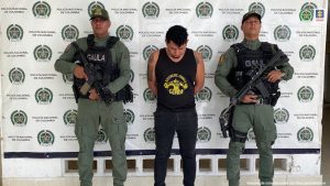 en la imagen se ve una persona detenida bajo custodia de dos integrantes de la Policía Nacional. Detrás suyo un backing institucional. 
