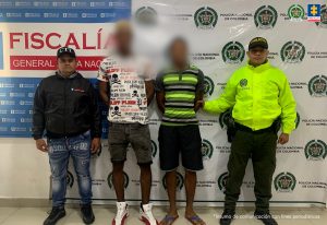 En la imagen se ven dos personas detenidas bajo custodia de un investigador del CTI y un integrante de la Policía Nacional. Detrás suyo backings institucionales.