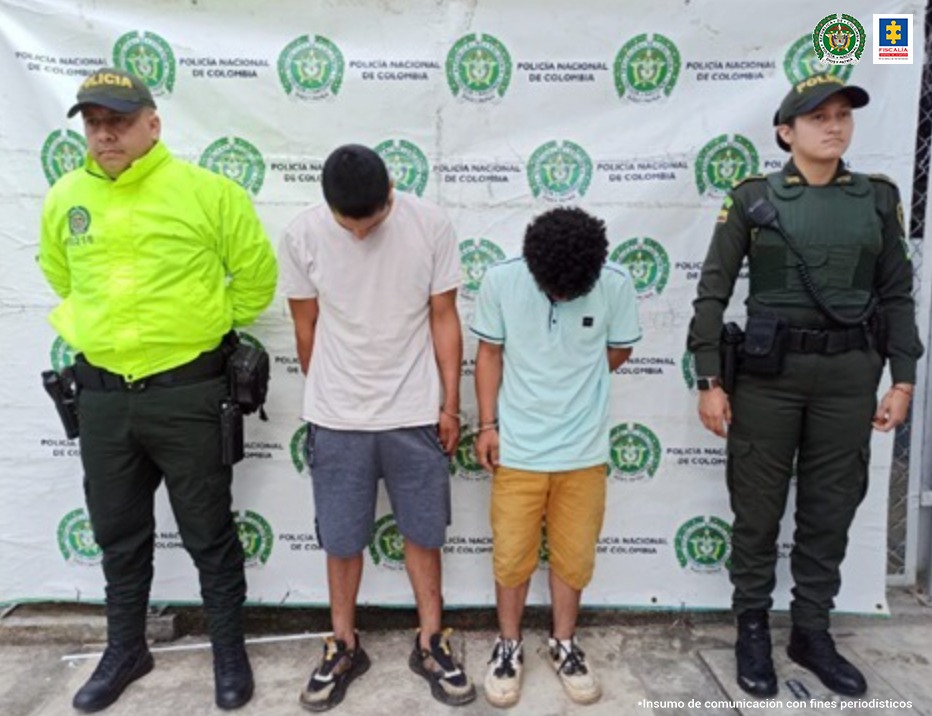 En la imagen se ven dos hombres detenidos bajo la custodia de dos miembros de la Policía Nacional. Detrás suyo un backing de la institución.
