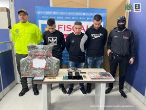 en la imagen se ven tres personas detenidas bajo custodia del CTI y la Policía Nacional. Frente a ellos varios elementos incautados sobre una mesa, detrás suyo backings institucionales.