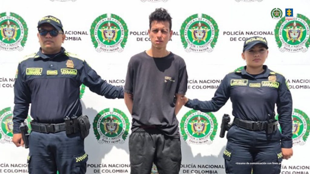 En la imagen se ve un hombre detenido bajo custodia de dos uniformados de la Policía Nacional. Detrás suyo, un backing institucional. 