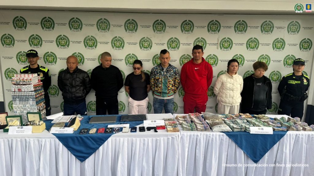 En la imagen se observa a ocho personas detenidas de pie frente a un fondo institucional de la Policía Nacional de Colombia, custodiadas por uniformados a ambos lados. Delante de ellas hay una mesa cubierta con un mantel blanco y azul sobre la que se exhiben fajos de dinero en efectivo, cajas de cigarrillos, documentos, teléfonos móviles, relojes y otros elementos incautados. La escena corresponde a un operativo de las autoridades contra una red de contrabando y lavado de activos.