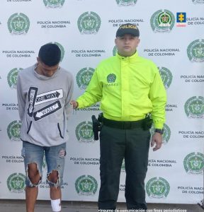 En la imagen se ve una persona detenida bajo custodia de un integrante de la Policía Nacional. Detrás suyo un backing institucional.