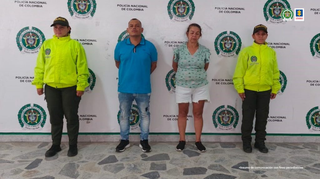 en la imagen se ve una pareja detenida bajo custodia de igual número de integrantes de la Policía Nacional. Detrás suyo un backing institucional.