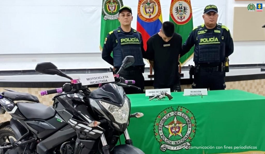 El procesado aparece esposado, custodiado por uniformados de la policía y está la moto incautada, así como el arma de fuego sobre una mesa.