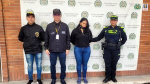 En la imagen se ve una mujer detenida bajo custodia de funcionarios del CTI, Policía Nacional y Gaula Militar. Detrás suyo un backing de la Policía.