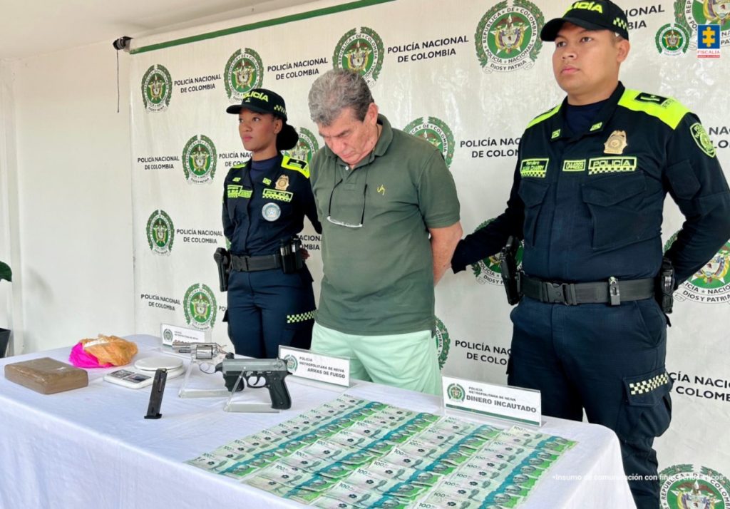 en la imagen se ve una persona detenida bajo custodia de dos uniformados de la Policía Nacional. Delante suyo una mesa con elementos incautados, detrás un backing institucional.