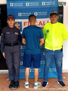 En la foto aparece de espaldas un hombre capturado, custodiado por una investigadora del CTI de la Fiscalía y un uniformado de la Policía Nacional. Las tres personas están delante de un banner de la Fiscalía.