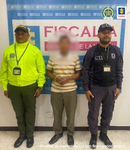 capturado de frente esposado manos adelante, custodiado por un policía y un investigador del CTI de la Fiscalía. Detrás banner de Fiscalía.