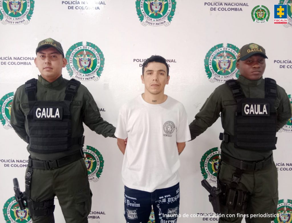 En la imagen se ve un hombre detenido bajo custodia de dos uniformados de la Policía Nacional. Detrás suyo un backing institucional.