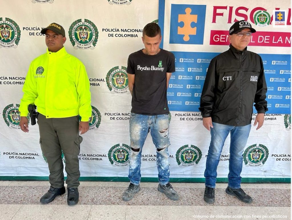 en la imagen se ve una persona detenida bajo custodia de un investigador del CTI y un integrante de la Policía Nacional. Detrás suyo un backing de cada una de las instituciones.