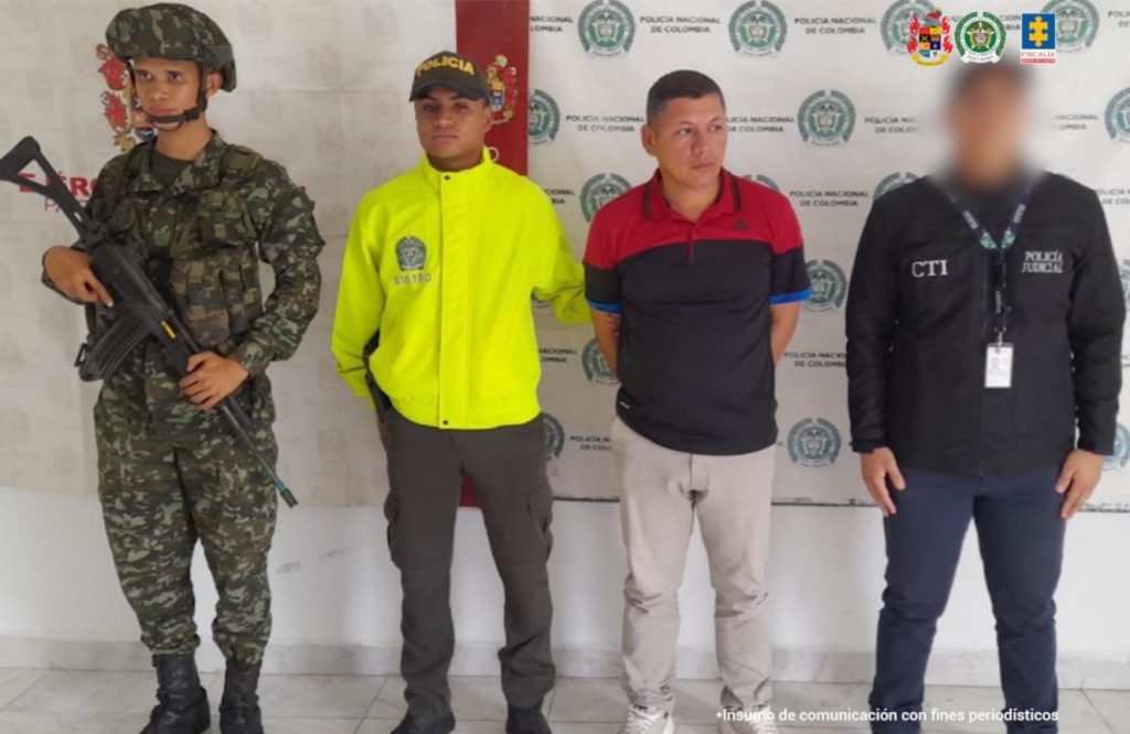 en la imagen se ve un hombre detenido bajo custodia de dos integrantes de la Policía Nacional. Detrás suyo un backing institucional.