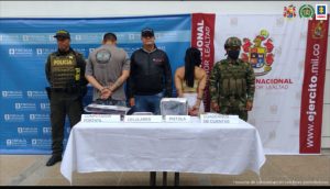 En la imagen se ven dos personas detenidas bajo custodia de un investigador del CTI, uno de la Policía Nacional y un integrante del Ejército. Detrás backings institucionales. Adelante, una mesa con elementos incautados entre ellos una pistola, celulares y otros.