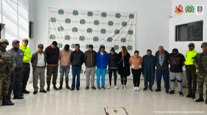 en la imagen se ven 13 personas bajo custodia de integrantes de la Policía y el Ejército, detrás suyo un backing institucional.