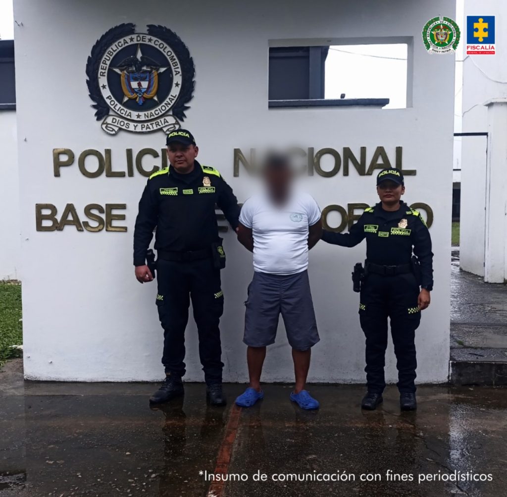El imputado quien tiene una pantaloneta y un buso blanco, se encuentra en custodia por dos uniformados de la Policía Nacional. Detrás se observa un letrero de la Policía.