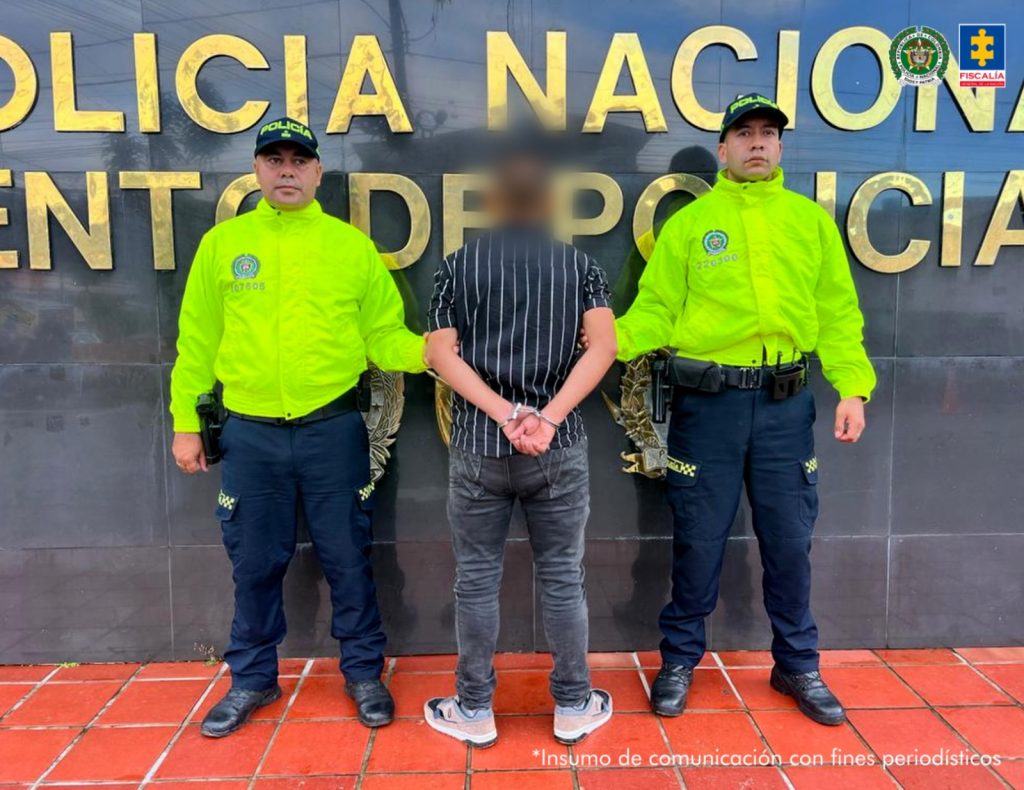 El imputado, se encuentra de espaldas con jean y buso negro de rayas y está en custodia por dos uniformados de la Policía Nacional que tienen una chaqueta verde. Detrás se observa un letrero de la Policía Nacional.
