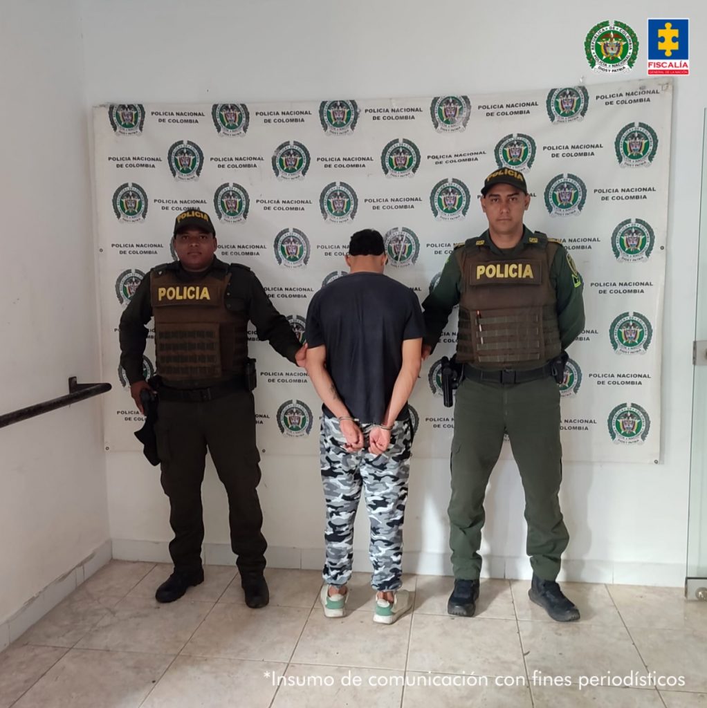 El imputado, se encuentra de espaldas con una sudadera camuflada y buso negro y está en custodia por dos uniformados de la Policía Nacional. Detrás se observa un letrero de la Policía Nacional.