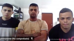 En la imagen aparecen tres hombres sentados, mirando de frente hacia la cámara.El hombre ubicado a la izquierda viste una camiseta negra con blanco y amarillo, con el logo de una marca de ropa visible. En el centro está un hombre con camiseta beige de manga corta, brazos cruzados y tatuajes en el antebrazo. A la derecha hay otro hombre con camiseta negra, que mantiene el rostro serio. El entorno corresponde a una habitación sencilla, con paredes blancas, una puerta marrón y un estante metálico con carpetas y libros al fondo. En la parte inferior de la imagen se alcanzan a ver textos superpuestos, entre ellos: “permanente (No comprobado)” y en la esquina inferior derecha la frase: “Insumo de comunicación con fines periodísticos”.