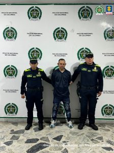 En la imagen se ve un hombre detenido bajo custodia de dos policías. Detrás un backing institucional.