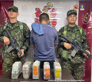 en la imagen se ve un hombre detenido y de espaldas, bajo custodia de dos militares. Delante de ellos una mesa con paquetes de dinero incautado.También le puede interesar: