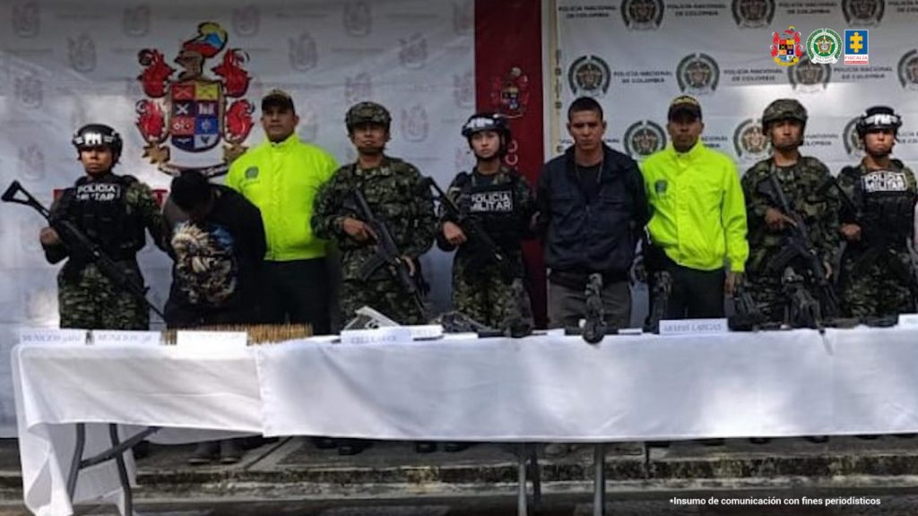En la imagen se ven dos personas detenidas, bajo custodia de integrantes de la Policía Nacional y el Ejército. Frente a ellos una mesa con elementos incautados. Detrás, un backing institucional.