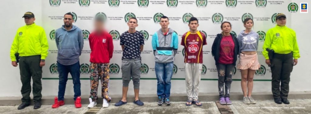 En la imagen se ve un grupo de personas detenidas bajo custodia de la Policía Nacional. Detrás suyo un backing de la misma institución.