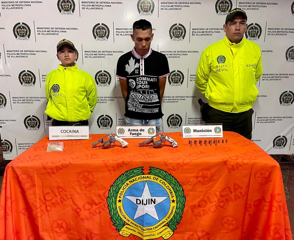 En la foto está en capturado de frente con las manos atrás, custodiado por un hombre y una mujer policías uniformados detrás está en banner de la policía y delante de ellos una mesa con el material incautado.