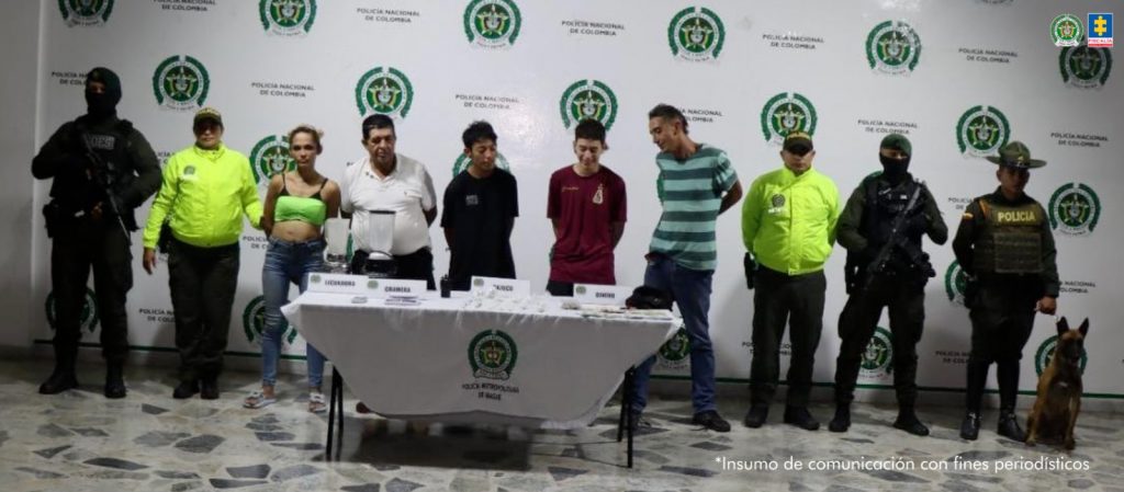 En la imagen se ven varias personas detenidas bajo custodia de integrantes de la Policía Nacional. Delante suyo una mesa con elementos incautados, detrás, un backing institucional.