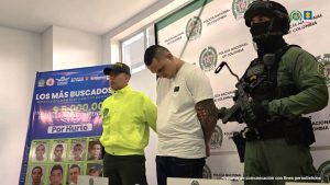 Los capturados están custodiados por uniformados de la Policía, y en una mesa frente a ellos, está un arma de fuego y equipos de comunicación incautados en los allanamientos.